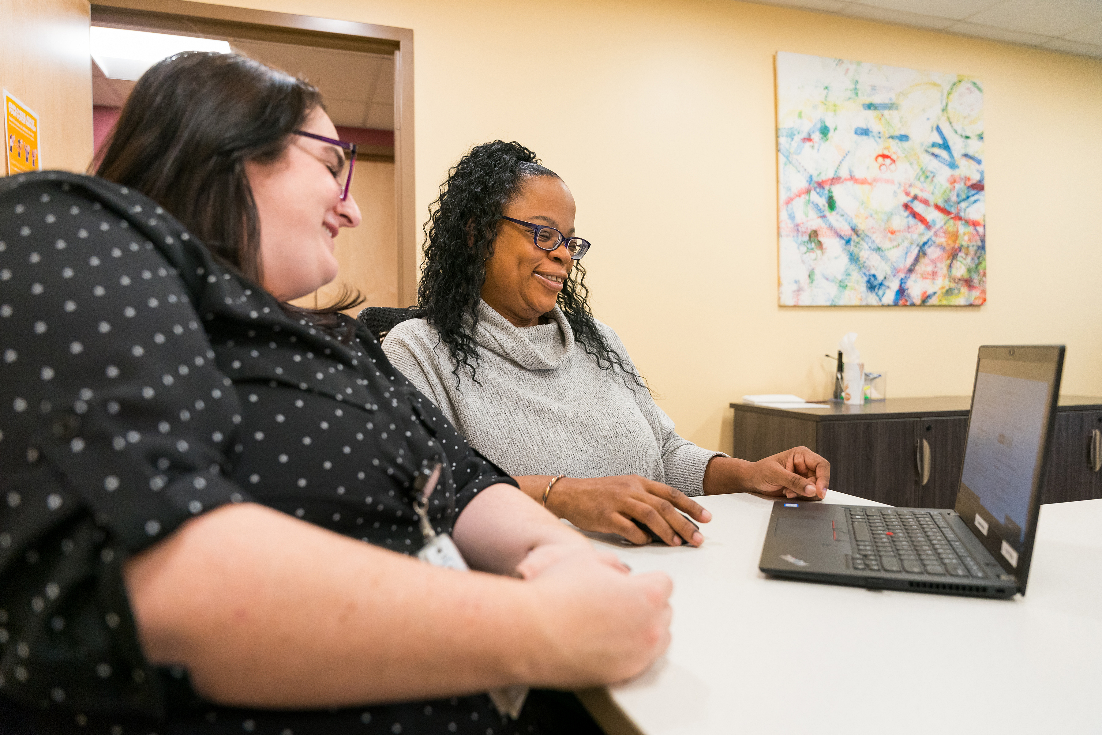 Two women smile while sitting at a table looking at a laptop computer.