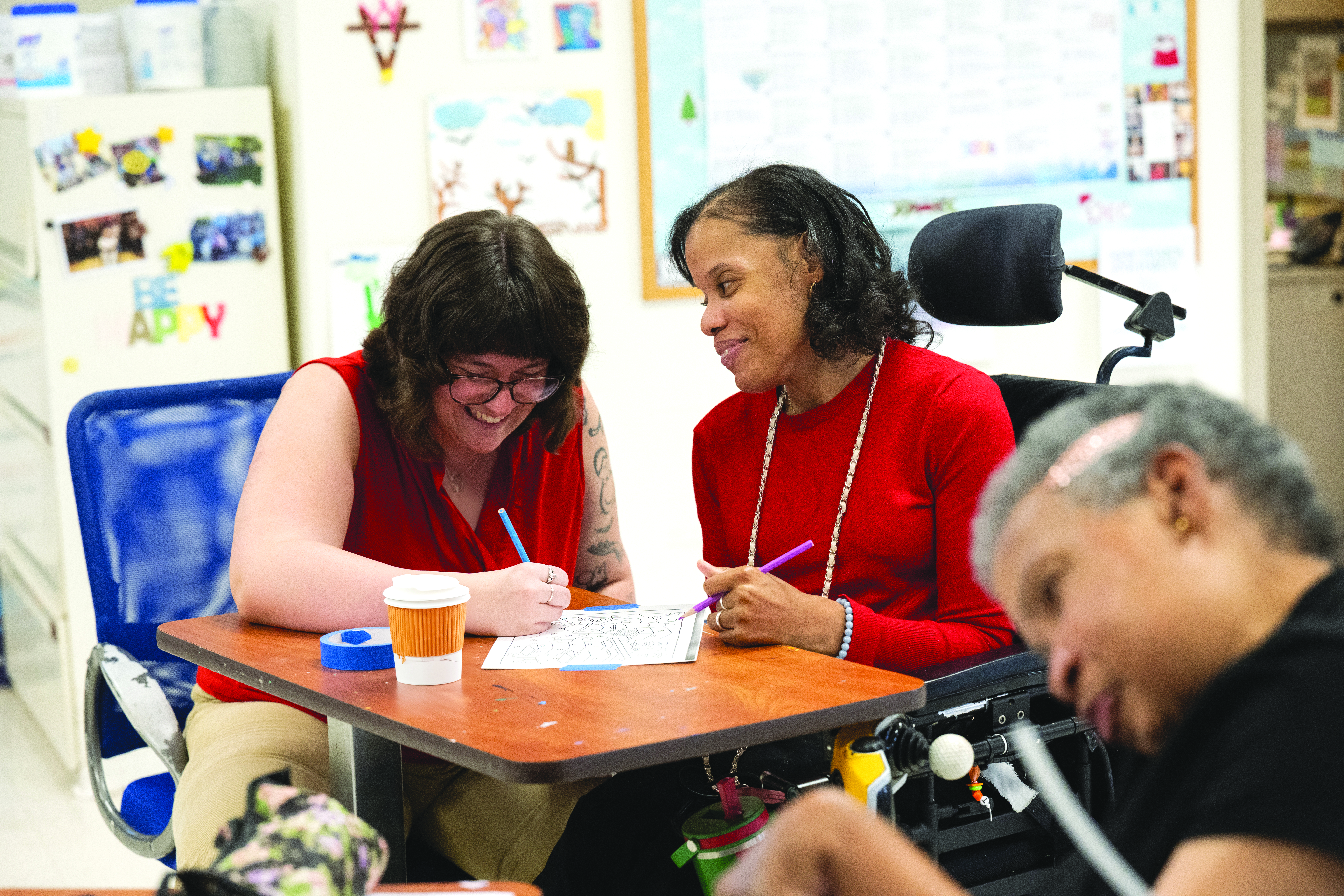 Two women wearing red, the one on the right using a wheelchair, work on an art project.