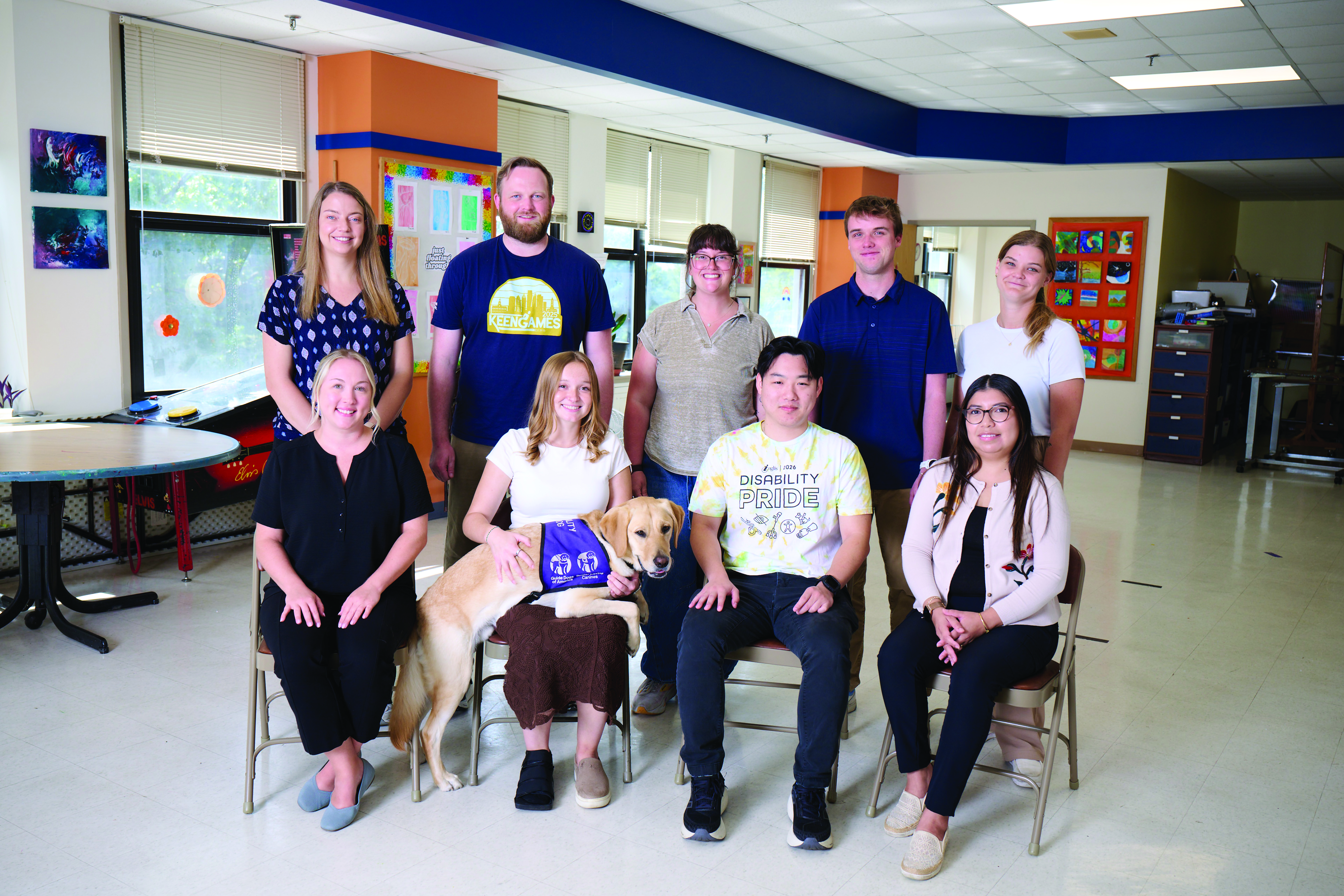 A group of people lined up in two rows, smiling for a picture, there's a dog in the lap of the 2nd person from the left in the first row.